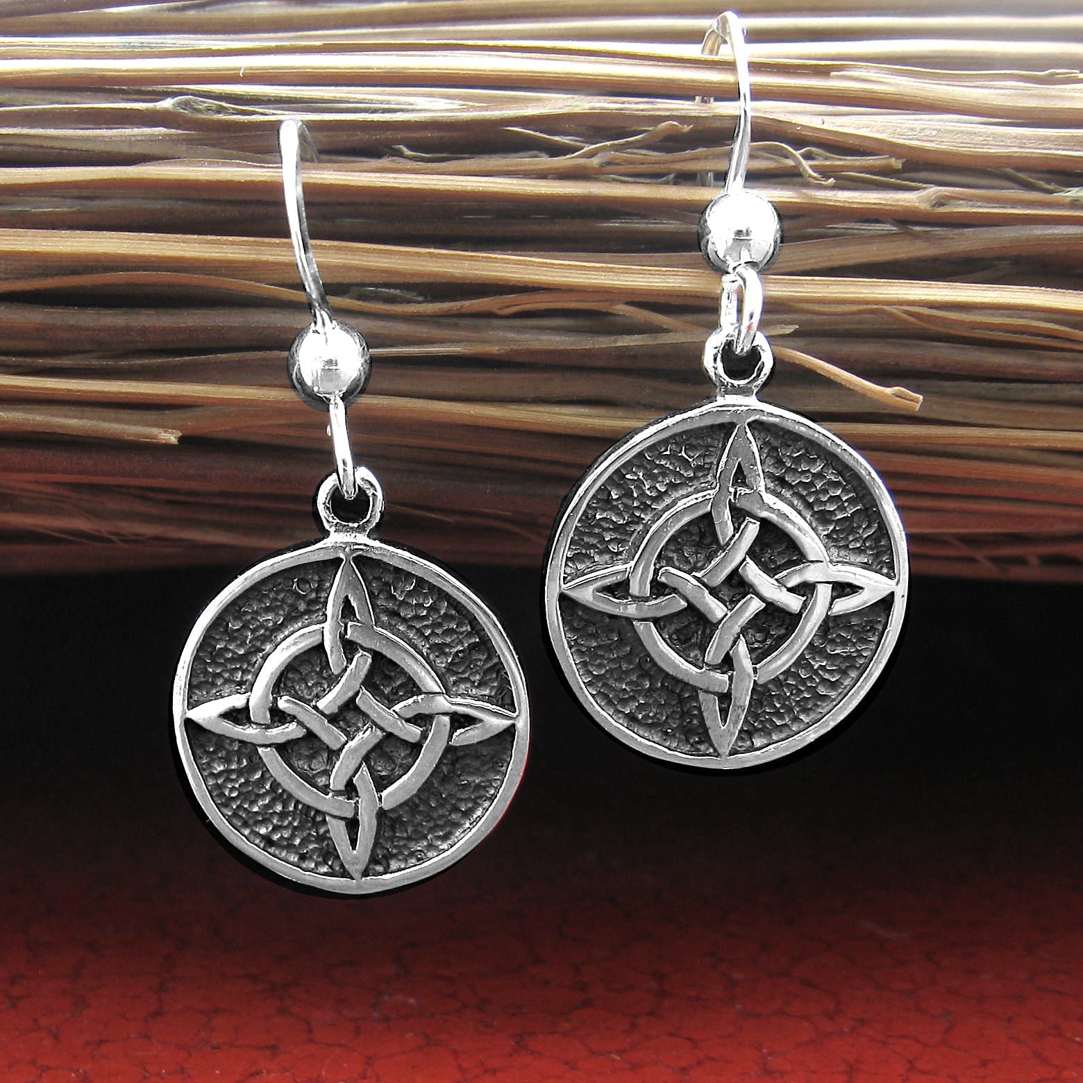 Two round silver hook earrings with oxidized Witch's Knot design hanging upright from the bristles of an old fashioned broom lying horizontally on a glazed red ceramic surface.