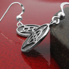 Side view of a round silver hook earring with oxidized Witch's Knot design leaning against a round dark wooden prop with a second round silver hook earring with oxidized Witch's Knot design lying flat on a glazed red ceramic surface.