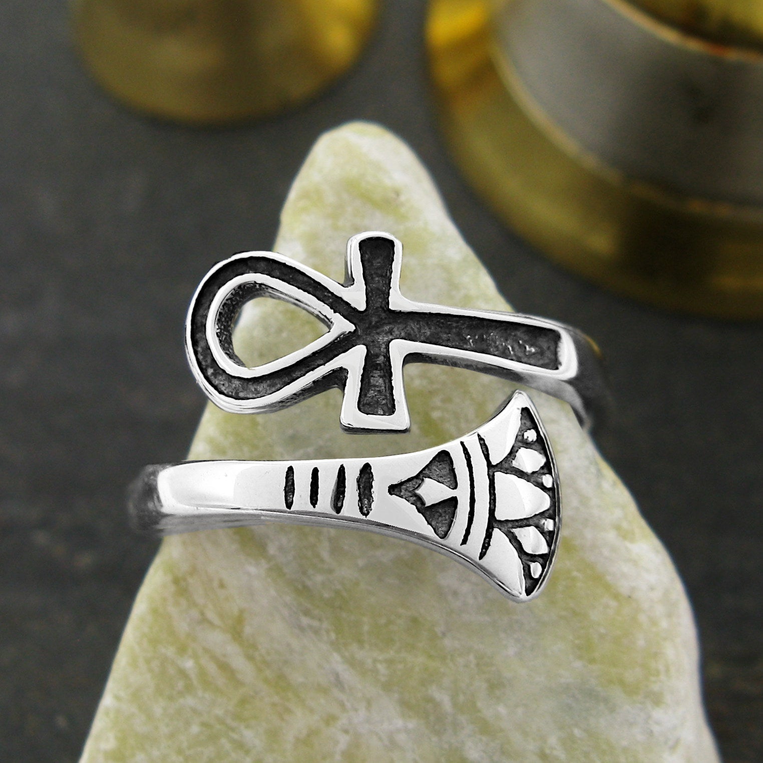 Front view of silver wraparound ring with ancient Egyptian Ankh symbol design with black accents displayed on triangular shaped light stone against out-of-focus dark brown and gold background.