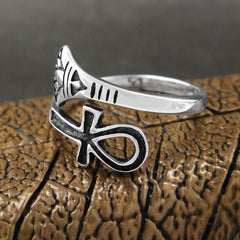 Front/side view of silver wraparound ring with ancient Egyptian Ankh symbol design with black accents sitting on shiny textured bronze-color metal surface against out-of-focus dark brown background.