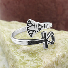 Front/side view of silver wraparound ring with ancient Egyptian Ankh symbol design with black accents sitting on flat light-colored stone against out-of-focus dark reddish background.