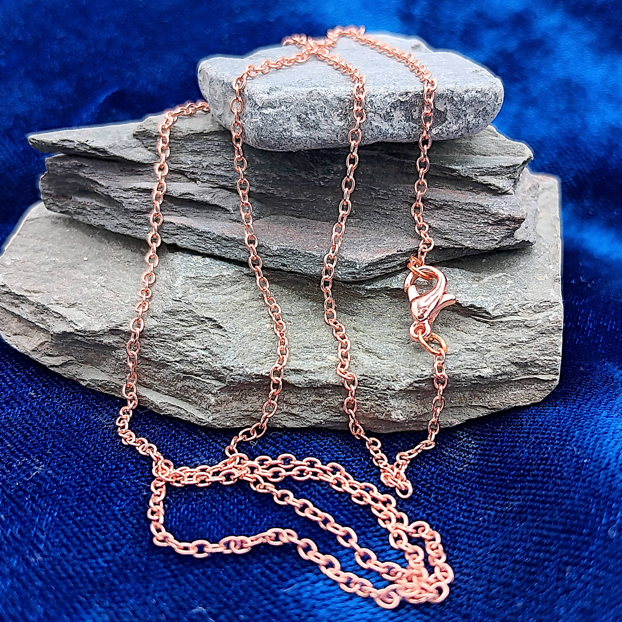 A delicate copper chain necklace draped over stacked gray stones on a textured blue fabric background.