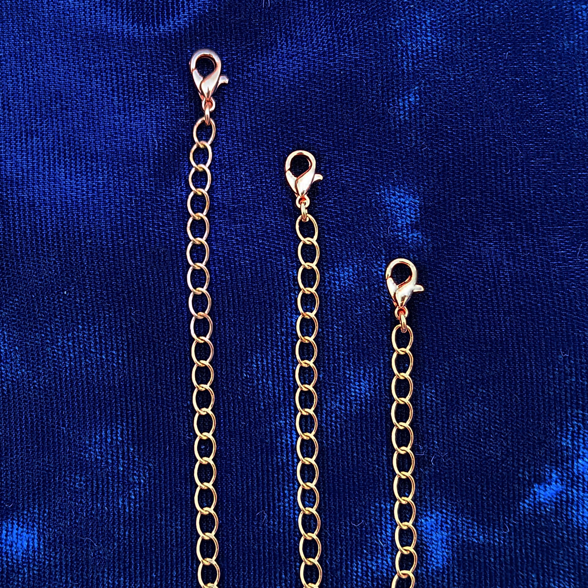 Three copper chain extender links with lobster clasps, displayed vertically against a textured blue velvet background.