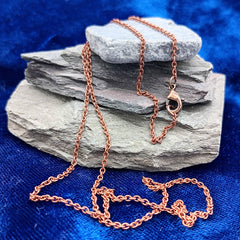A delicate copper chain necklace with a lobster clasp, draped over stacked gray slate stones on a textured blue fabric background.