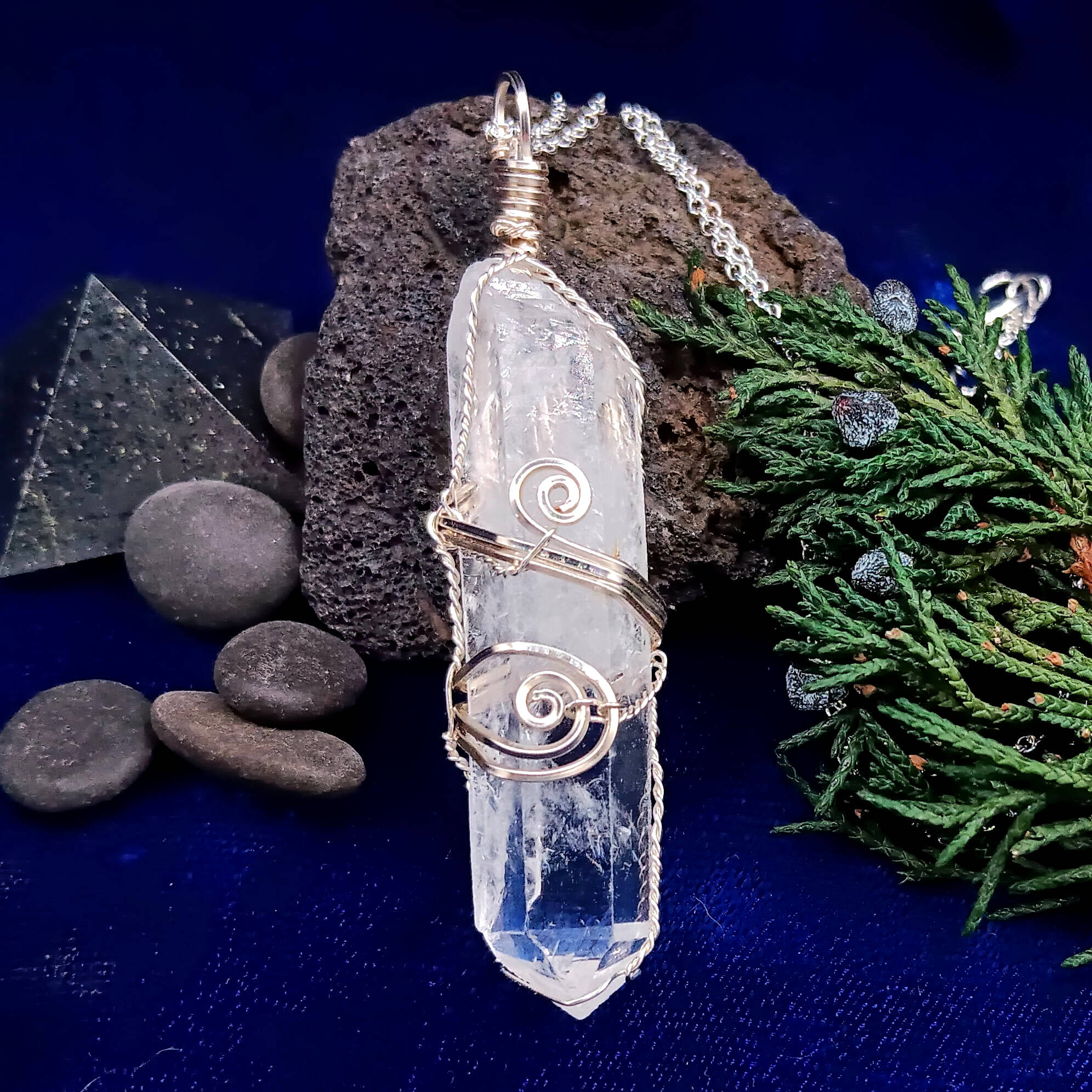 Crystal pendant with silver wire wrapping on a dark blue background with stones and greenery.