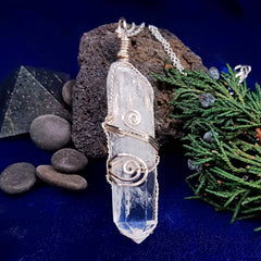 Crystal pendant with silver wire wrapping on a dark blue background with stones and greenery.