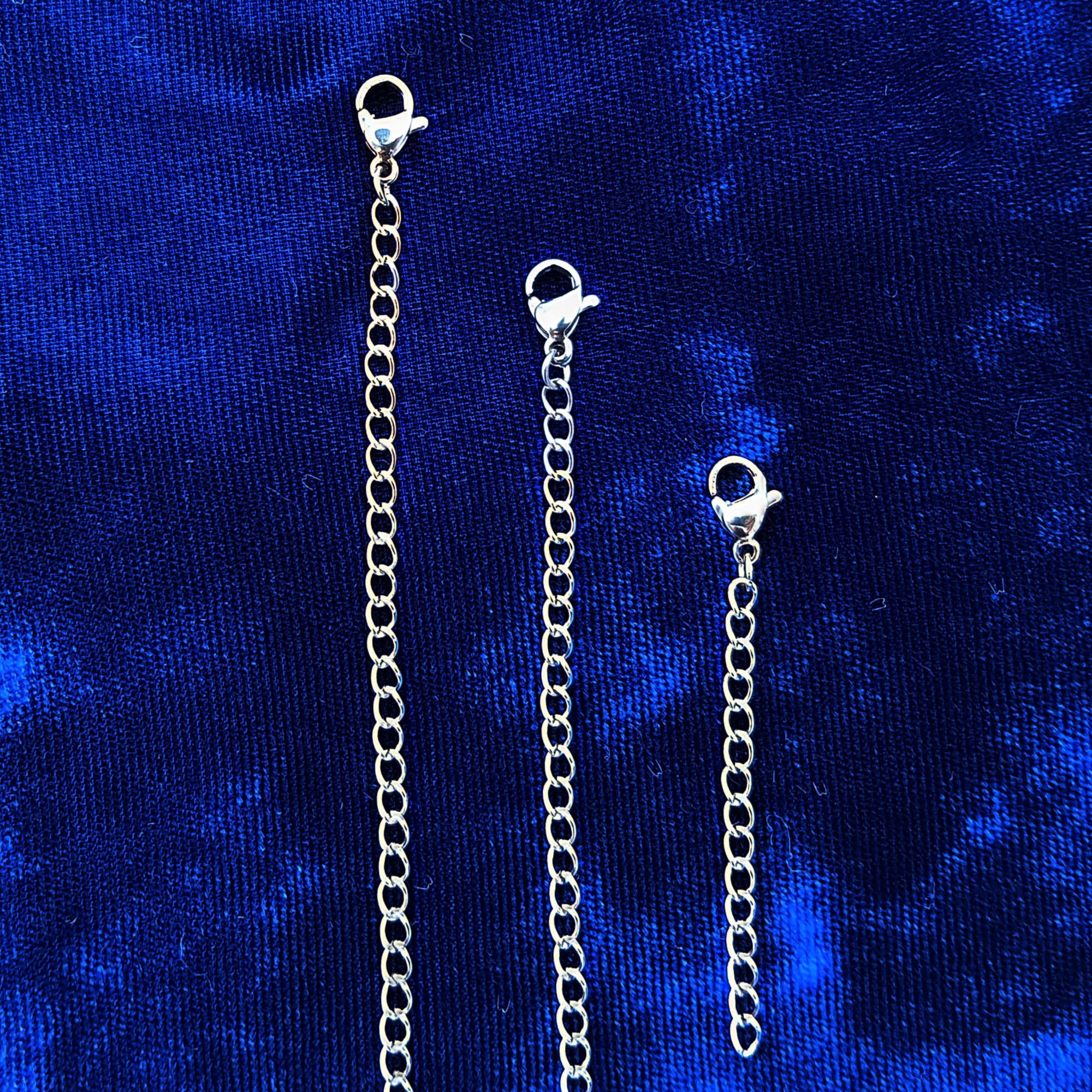 Three stainless steel chain extender links with lobster clasp ends, displayed vertically against a textured royal blue velvet background.