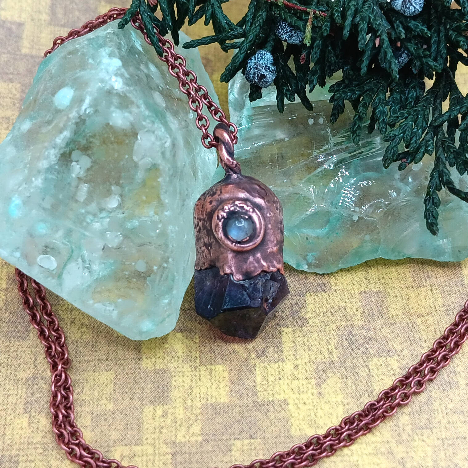 tiny raw amethyst crystal moonstone copper pendant against background of blue glass, mustard color paper, with chain
