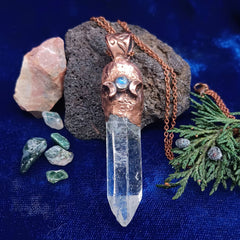 Copper pendant with triple moon and crystal on a blue fabric background with stones and greenery