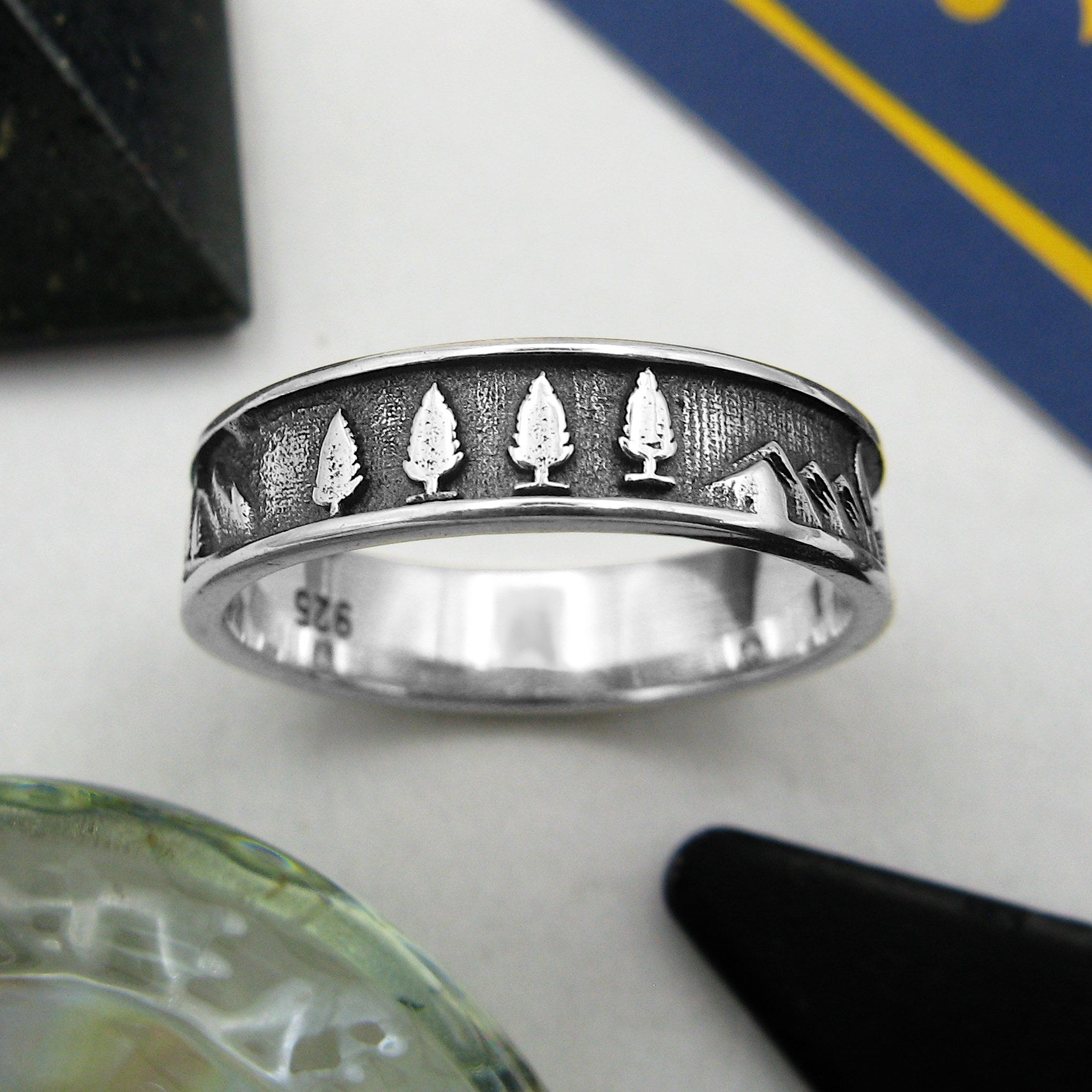 Oxidized sterling silver band type ring with trees and mountains design surrounded by various props on a white background.
