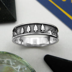 Oxidized sterling silver band type ring with trees and mountains design surrounded by various props on a white background.