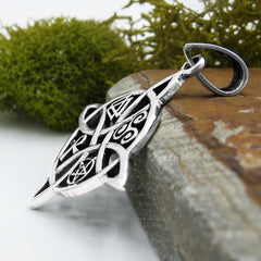 Side view of a silver witch's knot pendant with four symbols leaning against a stone terra cotta tile with green moss in the background and a white surface