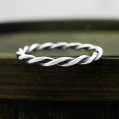 Slender silver ring with a braided rope pattern design sitting on the top edge of a dark wooden basket against a grayish background.