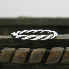 A twisted silver band ring, styled like a rustic rope, resting on a weathered wooden surface with a blurred grayish background.