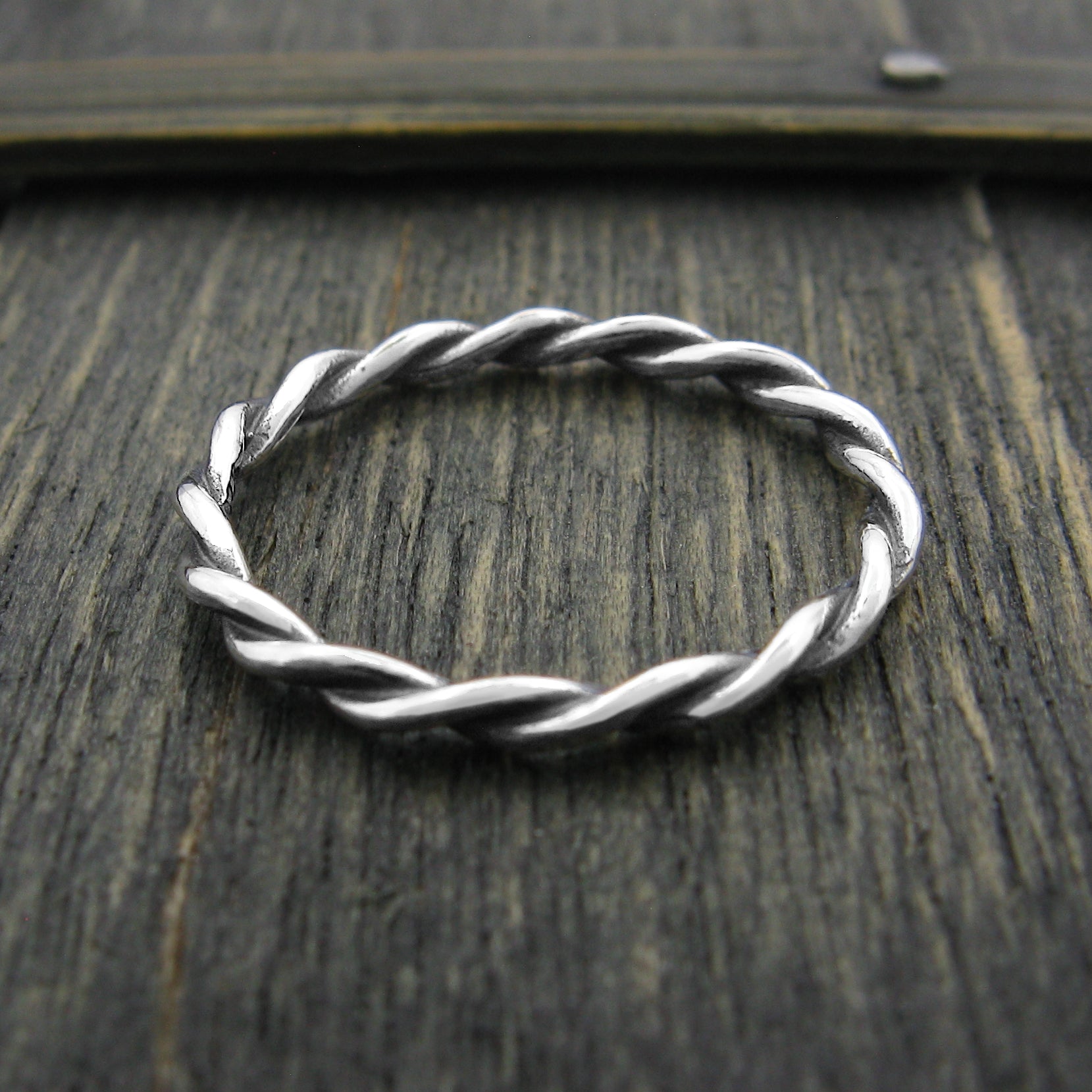 Overhead view of a slender silver ring with a braided rope pattern design sitting on the top edge of a dark greenish weathered wooden surface.