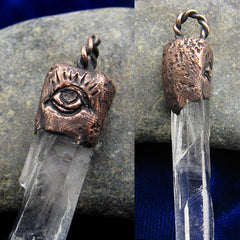 Close up detail view of top of pendant of clear quartz and textured copper cap with evil eye design and braided bail on smooth grey stone and dark blue cloth background.