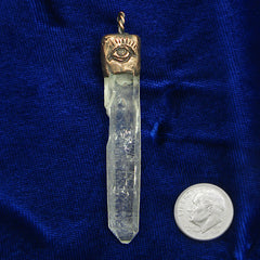 Pendant of clear quartz point with handmade textured copper cap with evil eye design and  braided copper bail next to a US dime on a dark blue cloth background.
