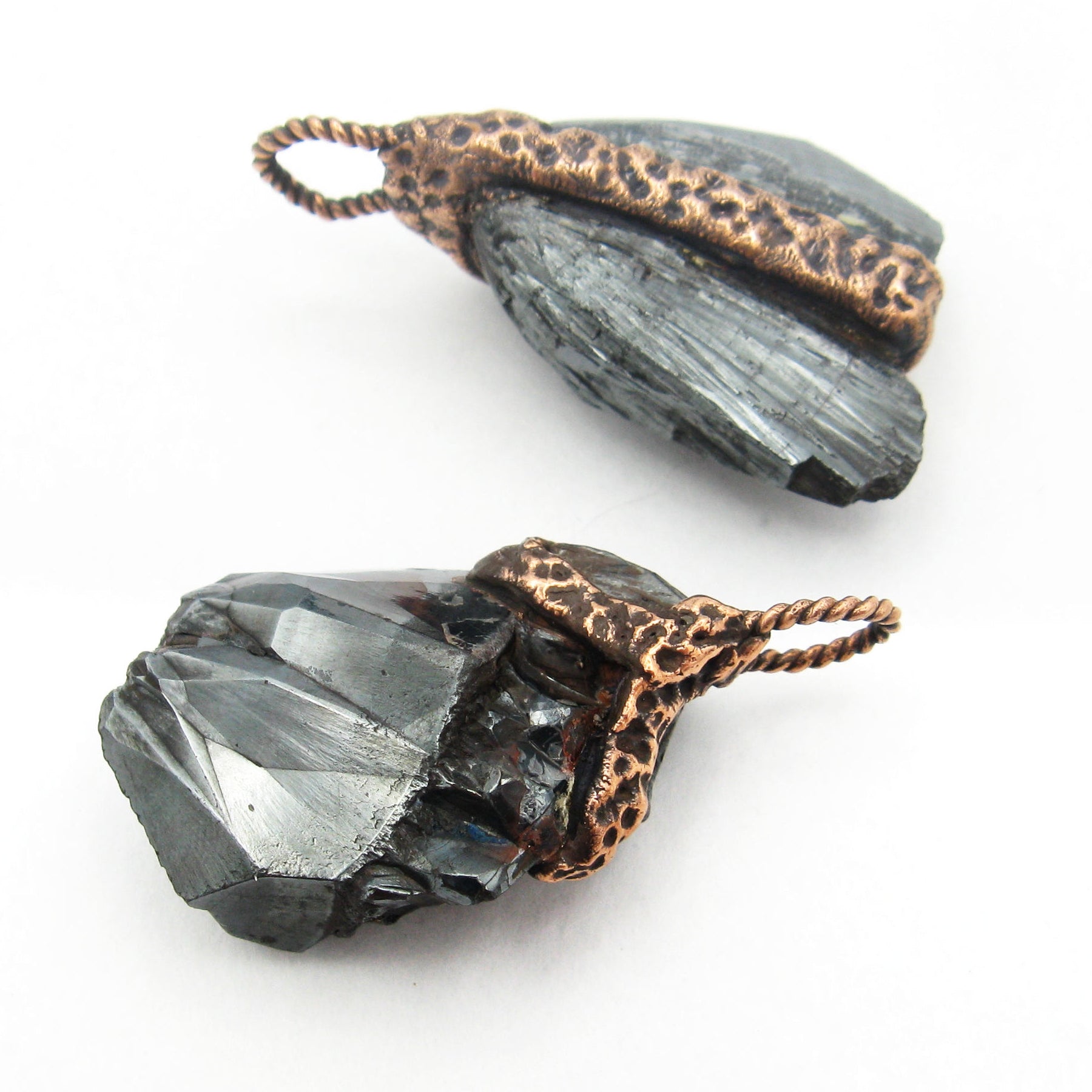 Side view of two randomly arranged handmade pendants made of hematite with textured copper harnesses and bails on white background.