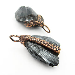Side view of two randomly arranged handmade pendants made of hematite with textured copper harnesses and bails on white background.