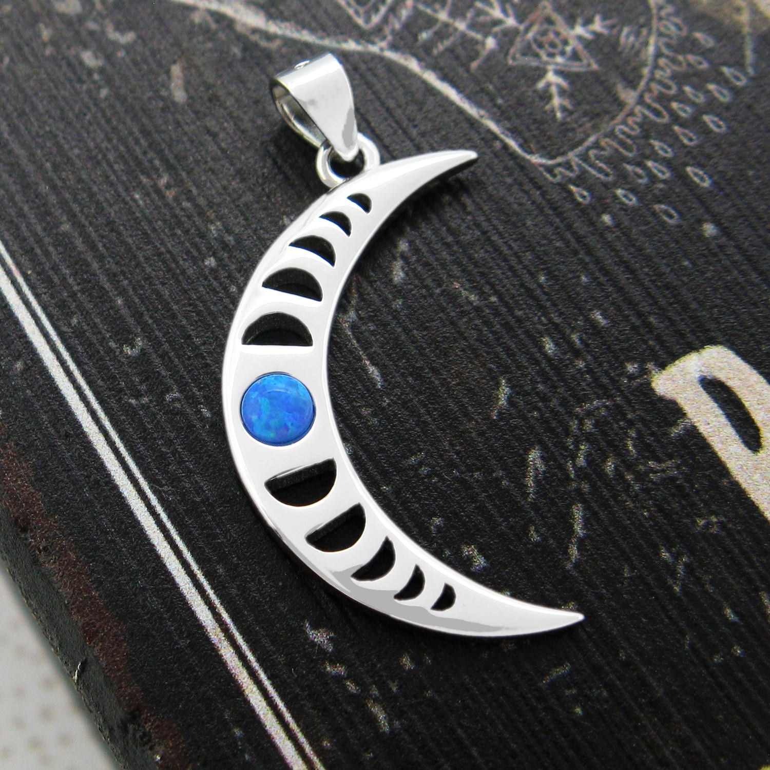 Sterling silver pendant in shape of crescent moon with blue cabochon and a cut-out moon-phases design on a weathered black wooden surface with white graphics.