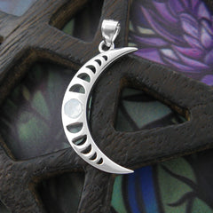 Sterling silver pendant in shape of crescent moon with moonstone cabochon and a cut-out moon-phases design on a weathered dark wooden pentagram prop on a colorful green and purple floral pattern background.