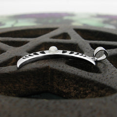 Side view of a sterling silver pendant in shape of crescent moon with a moonstone cabochon and a cut-out moon-phases design on a dark wooden pentagram prop on a blurry colorful green and purple floral pattern background.