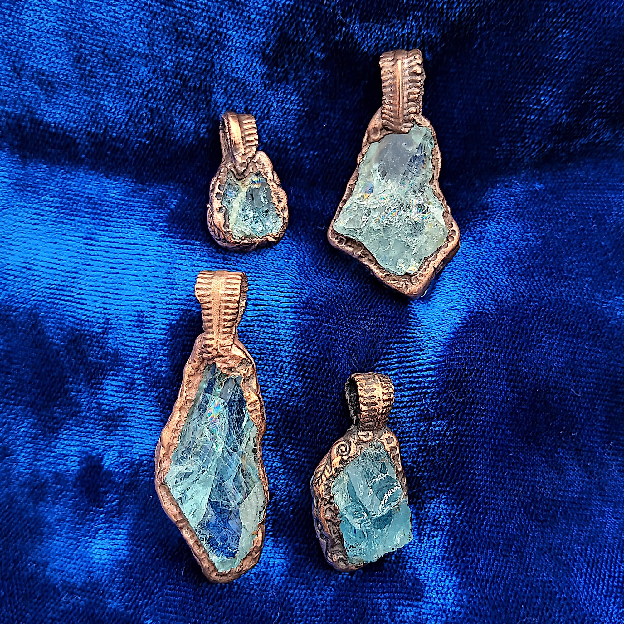 Four raw aquamarine crystal pendants with rough edges, set in textured copper, displayed on a vibrant blue velvet fabric background.
