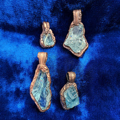 Four raw aquamarine crystal pendants with rough edges, set in textured copper, displayed on a vibrant blue velvet fabric background.